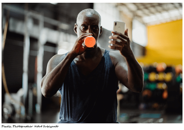 Black Male athlete drinking sports drink in gym while looking at his cell phone - selfie